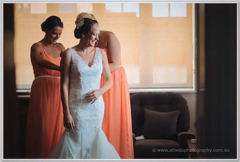 Bridesmaids in coral gowns clasp Katelyn's gown as she laughs in the sunlit bridal room