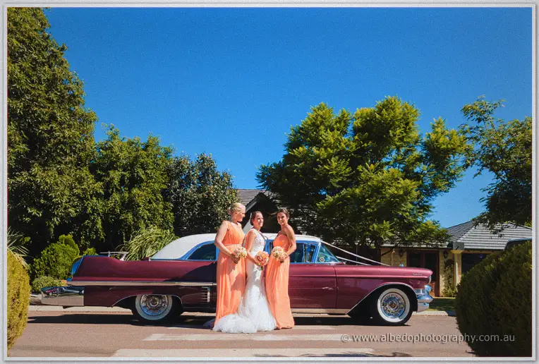 Katelyn and her two bridesmaids pose before the maroon Cadillac under a blue sky