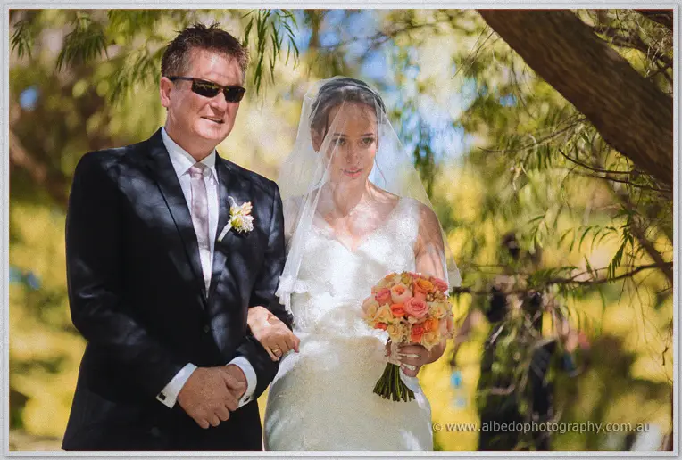 Katelyn walks the tree-lined aisle on her father's arm clutching peach roses