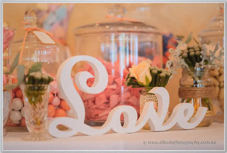 White wooden love sign surrounded by candy jars and peach roses on the dessert table