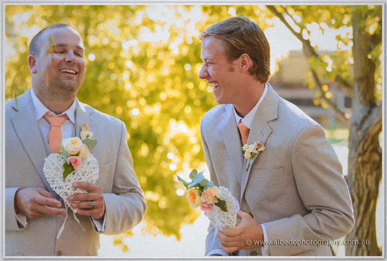 Groomsmen in taupe suits laughing beside golden foliage, each holding a white wicker heart with peach flowers