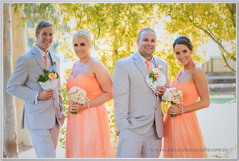 Bridal party smiling under a willow at Brookleigh, coral gowns and taupe suits with wicker heart bouquets