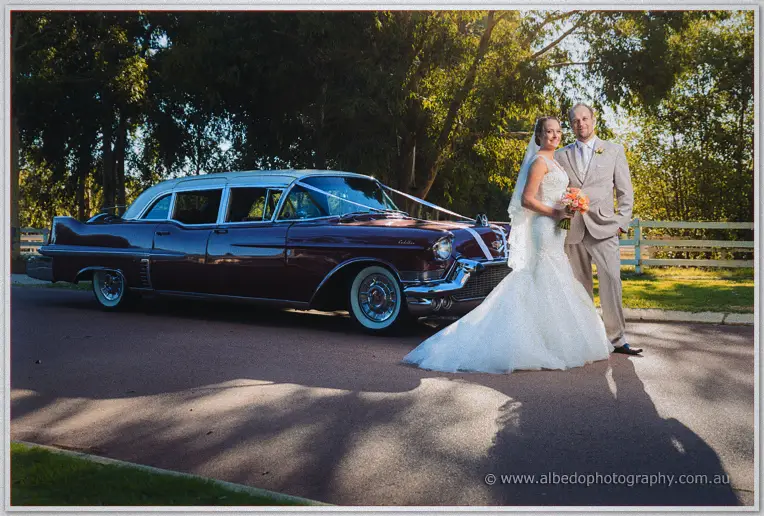 Katelyn and Matt beside their maroon vintage Cadillac on a tree-lined Brookleigh driveway in low sun