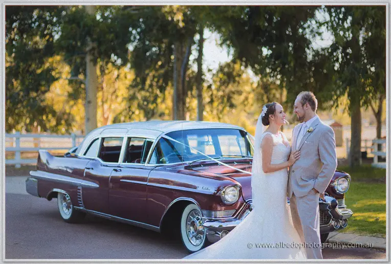 Katelyn and Matt smiling at each other beside the polished maroon Cadillac framed by sunlit trees