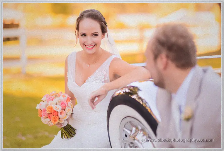 Katelyn laughing over her peach rose bouquet while leaning on the car wheel, Matt in soft-focus foreground