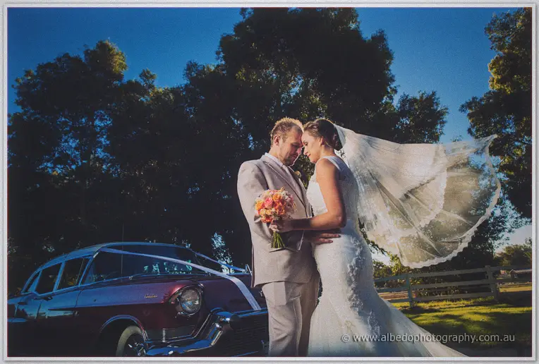 Matt and Katelyn forehead to forehead as her veil billows, vintage car and deep blue dusk sky behind