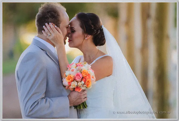 Katelyn cradling Matt's face as they kiss among the trees, peach rose bouquet between them