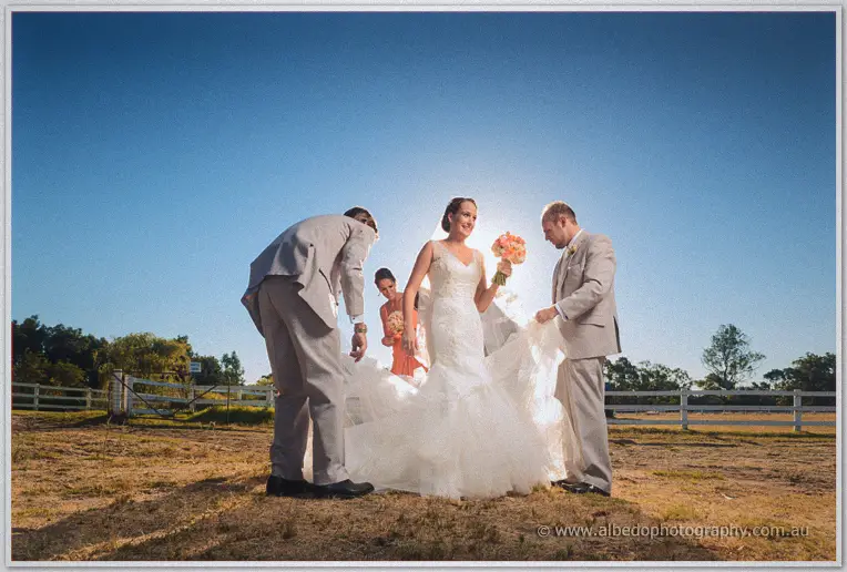 Bridal party lifting Katelyn's ruffled train in a dusty paddock, backlit by the sinking sun