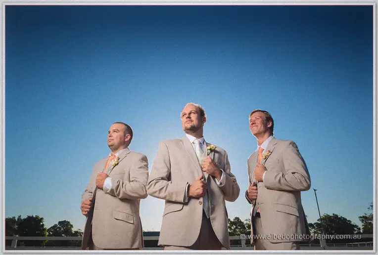 Low-angle portrait of Matt and his groomsmen buttoning taupe jackets against a deep blue sky