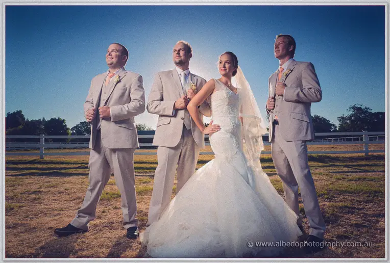 Katelyn posing with Matt and the groomsmen in golden paddock light, white fence stretching behind
