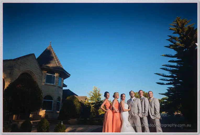Bridal party gathered on the lawn before the stone homestead's turret under a huge evening sky