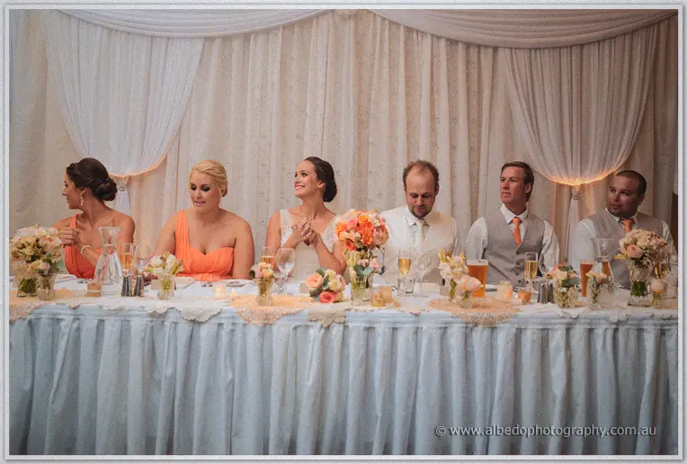 Bridal table mid-speech, Katelyn beaming between bridesmaids in orange and groomsmen in ties