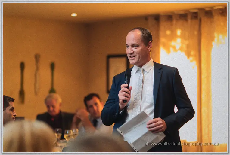 Groomsman in a navy suit delivering a toast with notes in hand, guests smiling behind