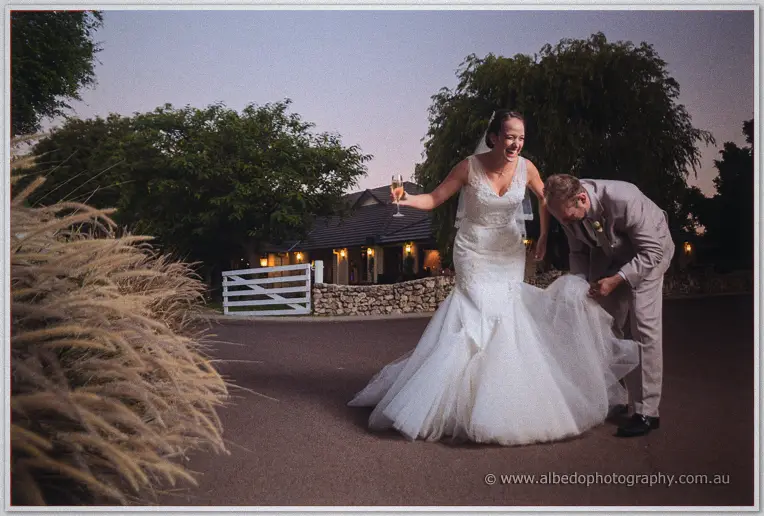 Katelyn laughs with a champagne glass while Matt gathers her gown train at dusk at Brookleigh