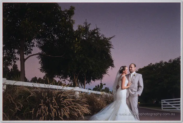 Katelyn and Matt embrace beneath a violet dusk sky by the white paddock fences of Brookleigh
