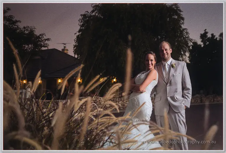 Katelyn and Matt smile arm in arm at dusk as golden grass plumes blur the foreground