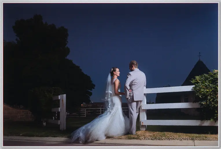 Katelyn and Matt lean together against a white ranch fence in the last light at Brookleigh
