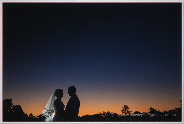 Katelyn and Matt stand forehead to forehead, silhouetted against a dusk sky fading to orange