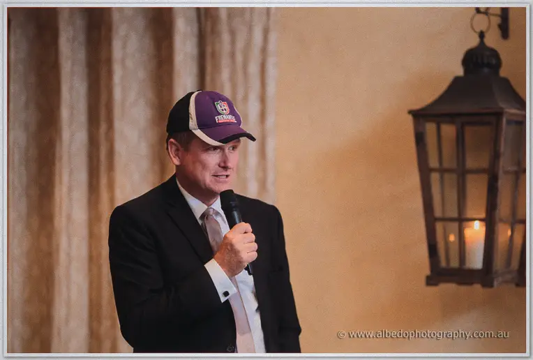 A groomsman in a purple cap gives a toast, microphone in hand, beside a wall lantern