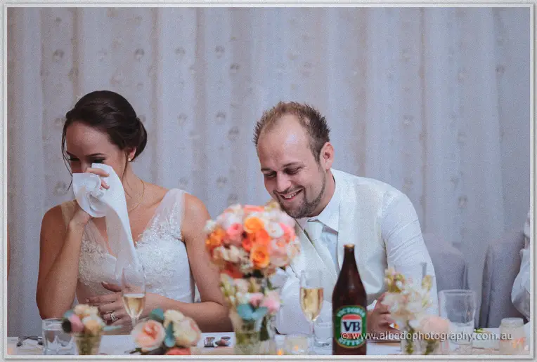 Katelyn wipes tears of laughter with a napkin while Matt grins during wedding speeches