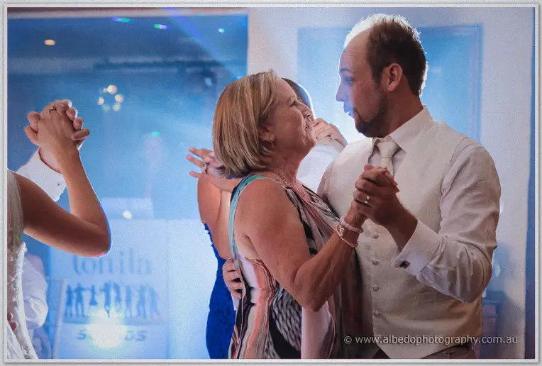 An older woman and a groomsman in a cream vest dance close under reception lights