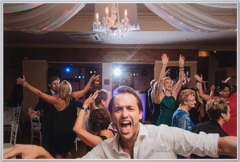 Guests throw their arms up beneath the chandelier while a man shouts with joy up front