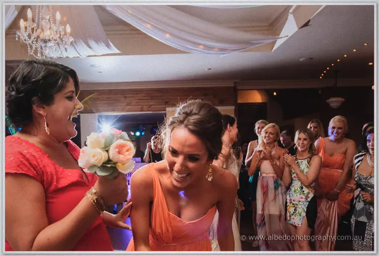 A guest in coral clutches the caught bouquet while friends cheer beneath the chandelier