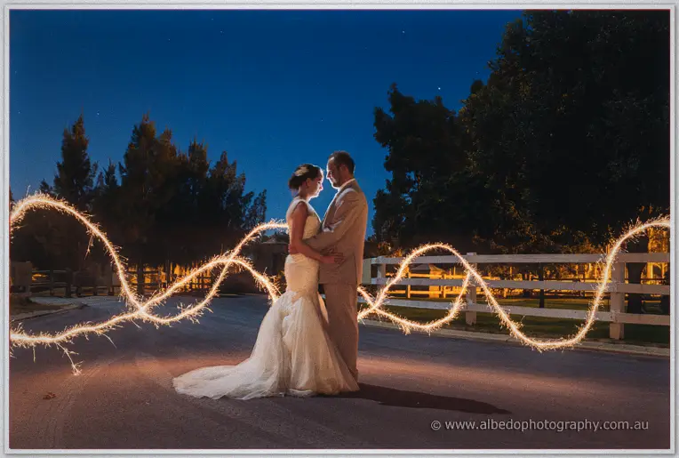 A swirling sparkler trail loops around Katelyn and Matt by the white fence at night