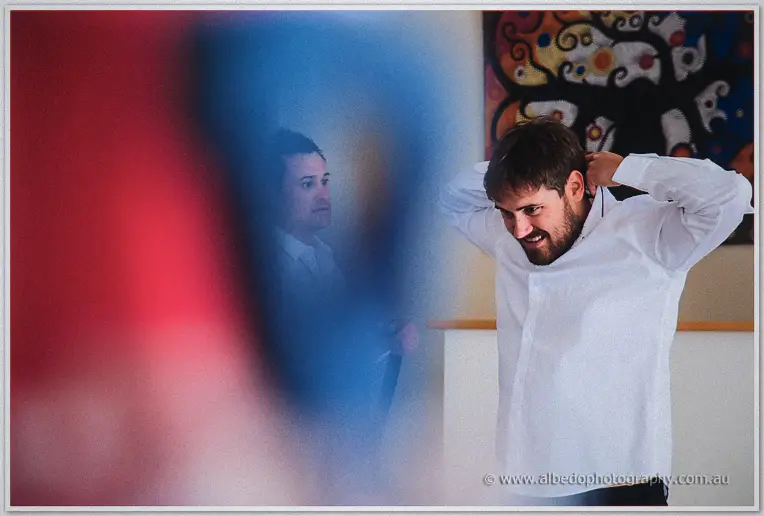 Bearded groomsman buttoning his collar in a mirror while the bride's family waits behind