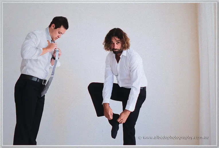 Groomsmen getting ready, one knotting his grey tie, a long-haired groomsman pulling on his sock