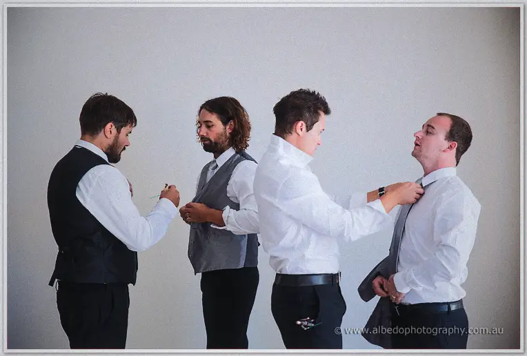 Four groomsmen in white shirts and grey vests helping each other with ties before the ceremony