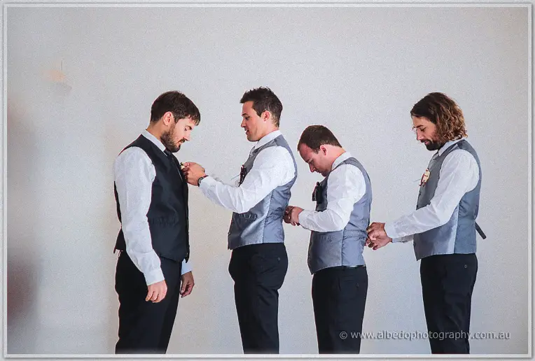Chain of groomsmen fastening vests and boutonnieres in a line against the white wall