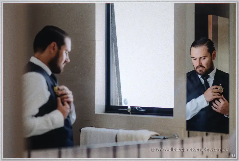Groom pinning his boutonniere before the bathroom mirror in soft morning light