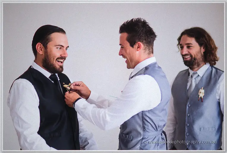 Groom laughing as a groomsman pins a small toy boutonniere to his black vest