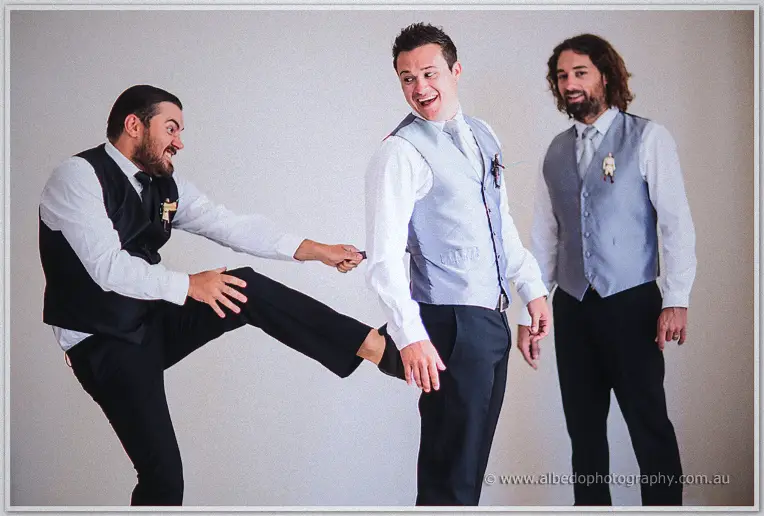 Groomsmen joking around, one kicking his leg high while the others laugh in their vests