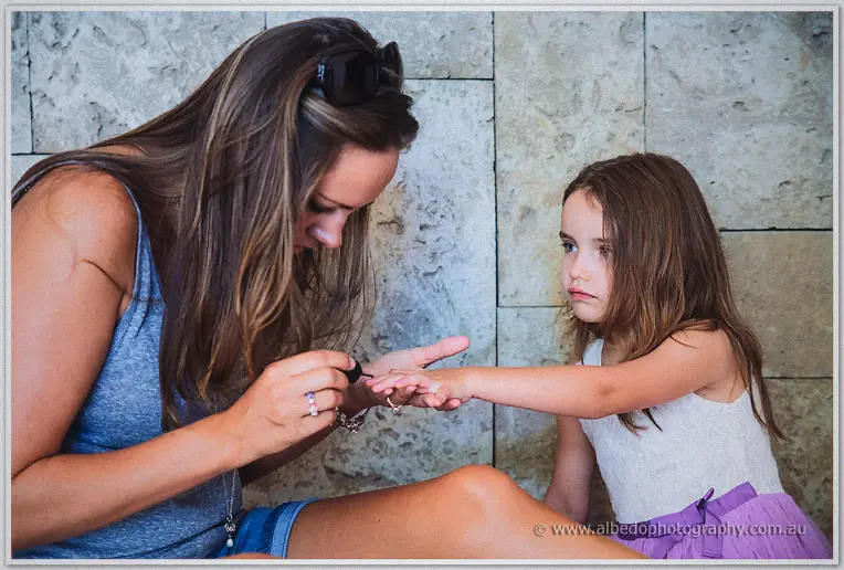 Woman painting a young girl's nails on a stone bench in the afternoon light