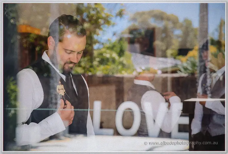 Groom's reflection in the poolside glass adjusting his toy boutonniere near the LOVE sign