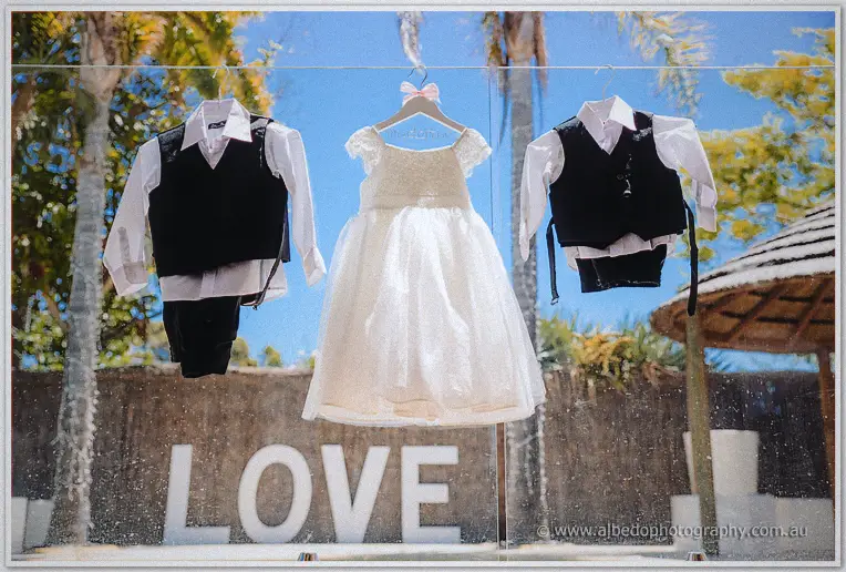 Flower girl dress and two page boy outfits hanging on the pool fence above white LOVE letters