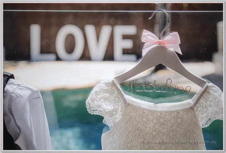 Lace flower girl dress on a hanger wired with the name Madeleine, pool and LOVE letters behind
