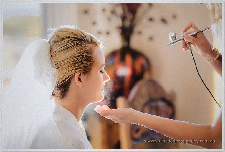 Makeup artist airbrushes Teigan's face as she sits veiled in soft window light before the ceremony