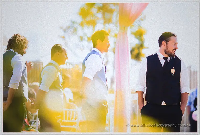 Troy grins among his groomsmen in golden backlight beneath the pink ceremony draping