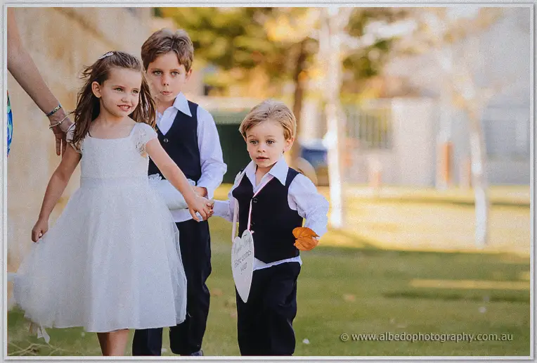 Two page boys in vests walk hand in hand with a flower girl across the lawn
