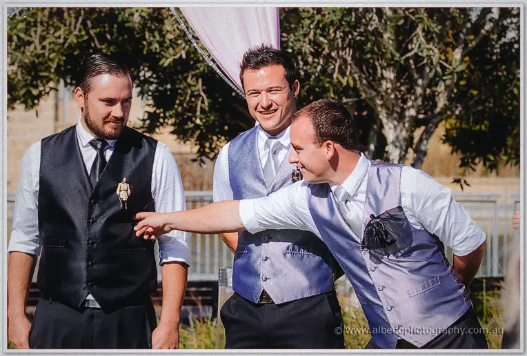 A groomsman reaches to pin Troy's boutonniere as the party laughs under the trees