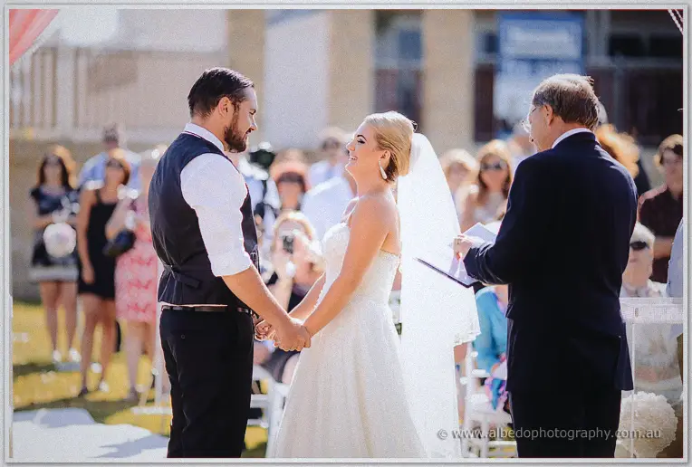 Troy and Teigan hold hands facing the celebrant with guests watching the garden ceremony