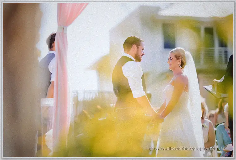 Troy and Teigan smile at each other through golden flare beneath the pink ceremony drape