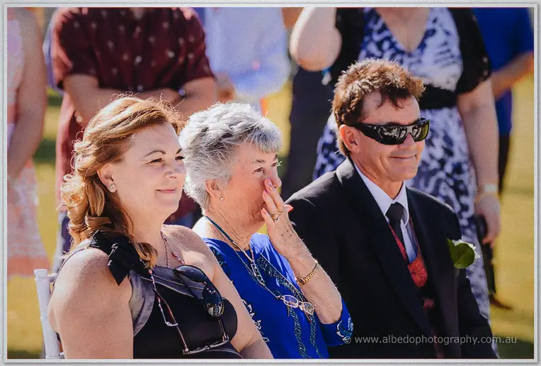 Teary wedding guests seated in the sun react as the couple exchange their vows
