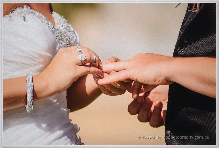 Close-up of Teigan's jewelled hands placing a wedding band on Troy's finger