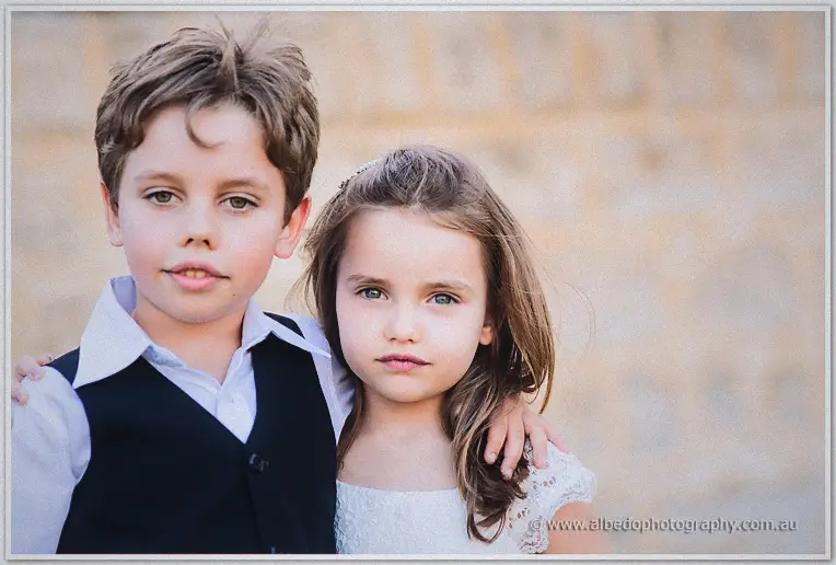 Page boy and flower girl pose solemnly together in soft light by the garden wall