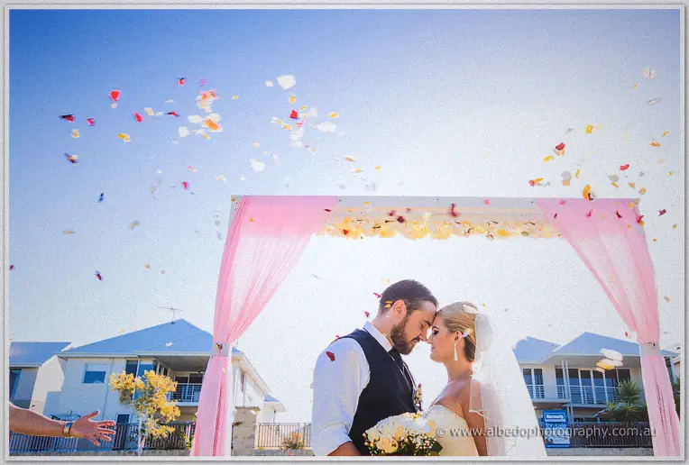 Petal confetti rains over Troy and Teigan beneath the pink arbour against blue sky