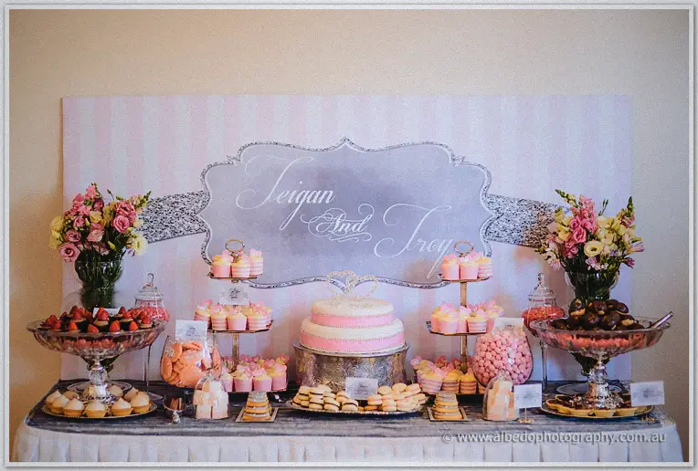 Teigan and Troy dessert bar with pink cake, cupcakes and lollies beneath their glittering sign